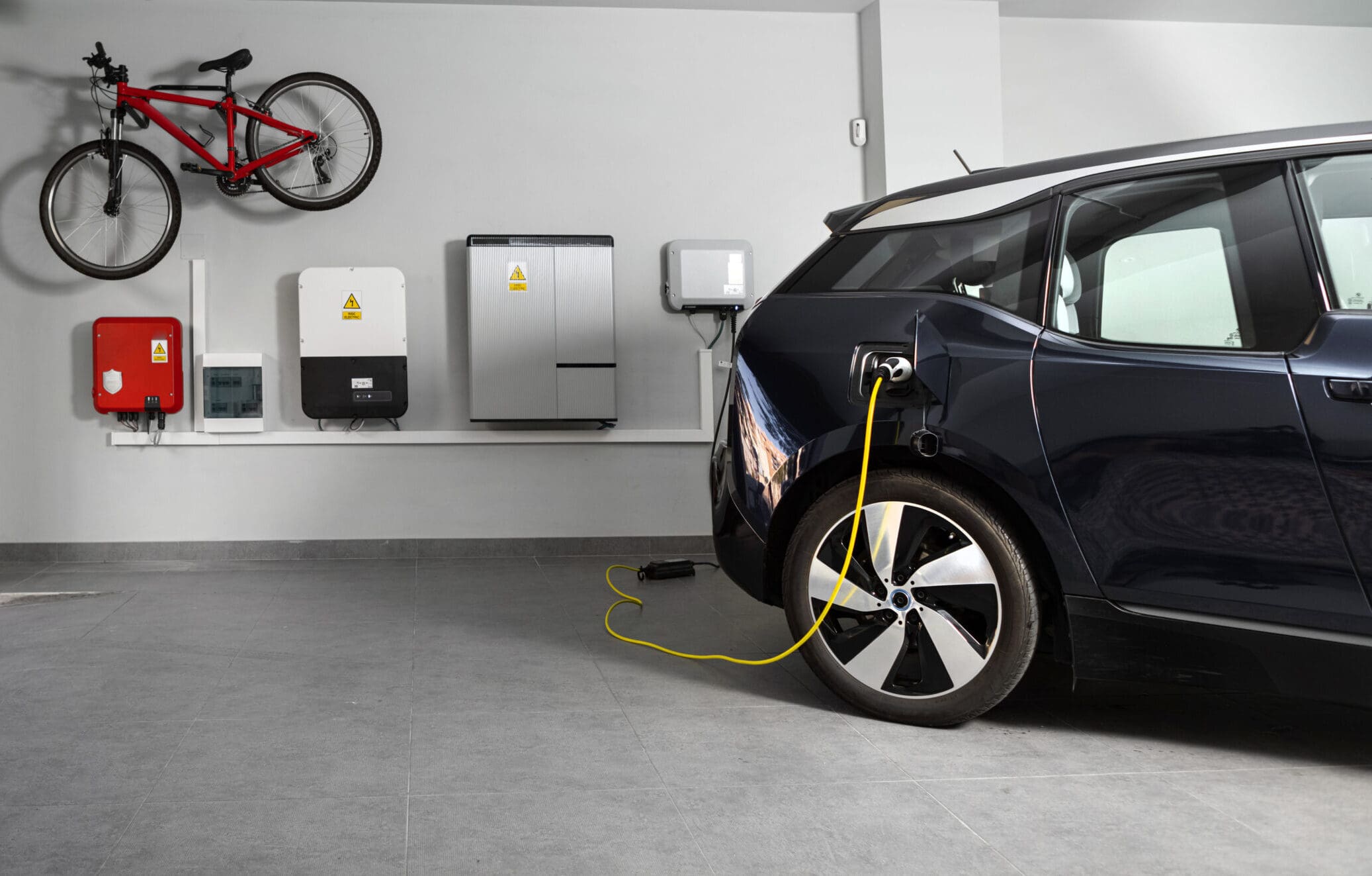 A black electric car is charging in a modern garage, highlighting recent electrical installations. A yellow cable connects to the car, with wall-mounted power units and a red bicycle displayed on the light gray wall above.