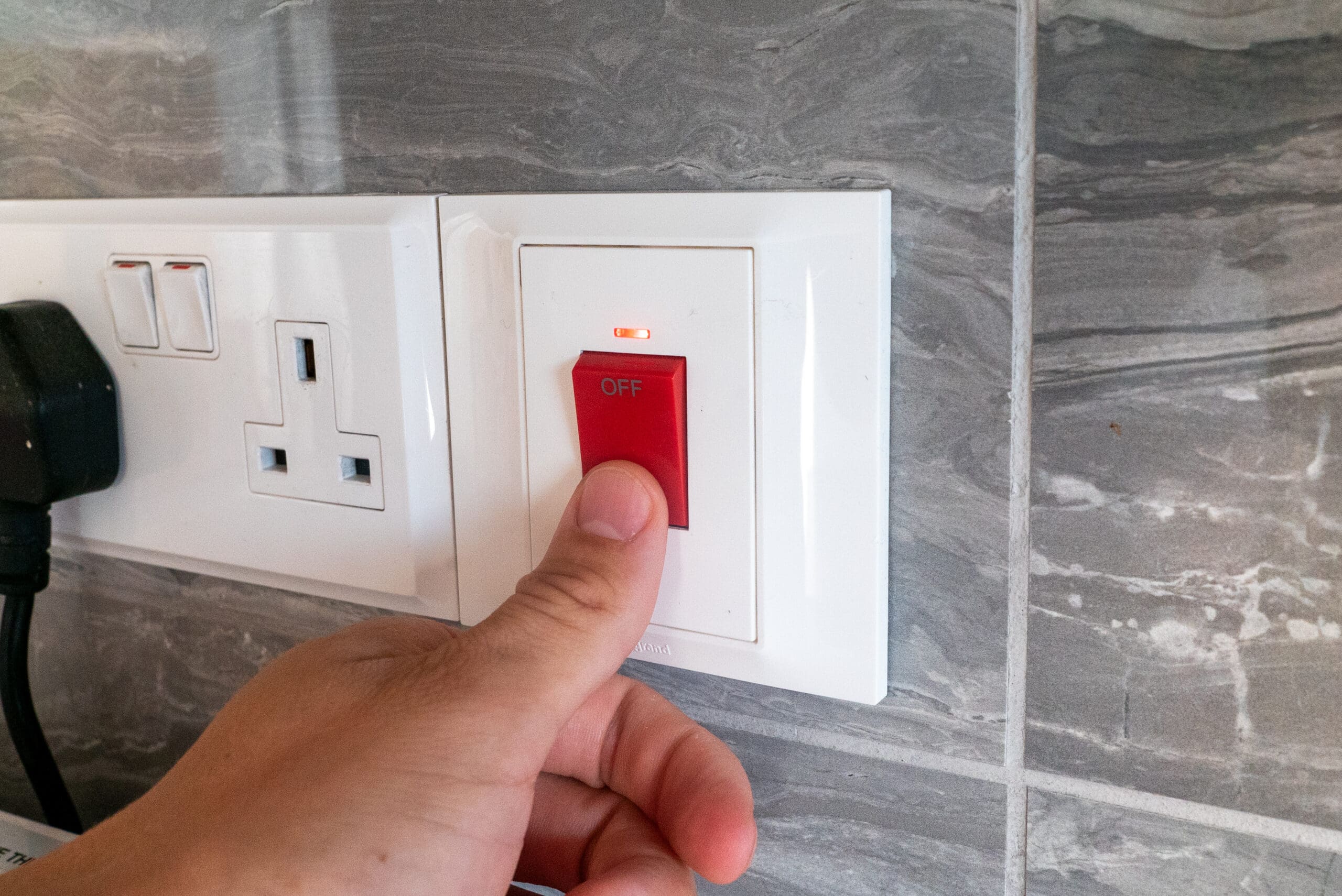 A hand is about to flip a red switch labeled "OFF" on a white power outlet panel, part of recent electrical installations, mounted on a tiled wall next to a plugged-in black electrical plug.