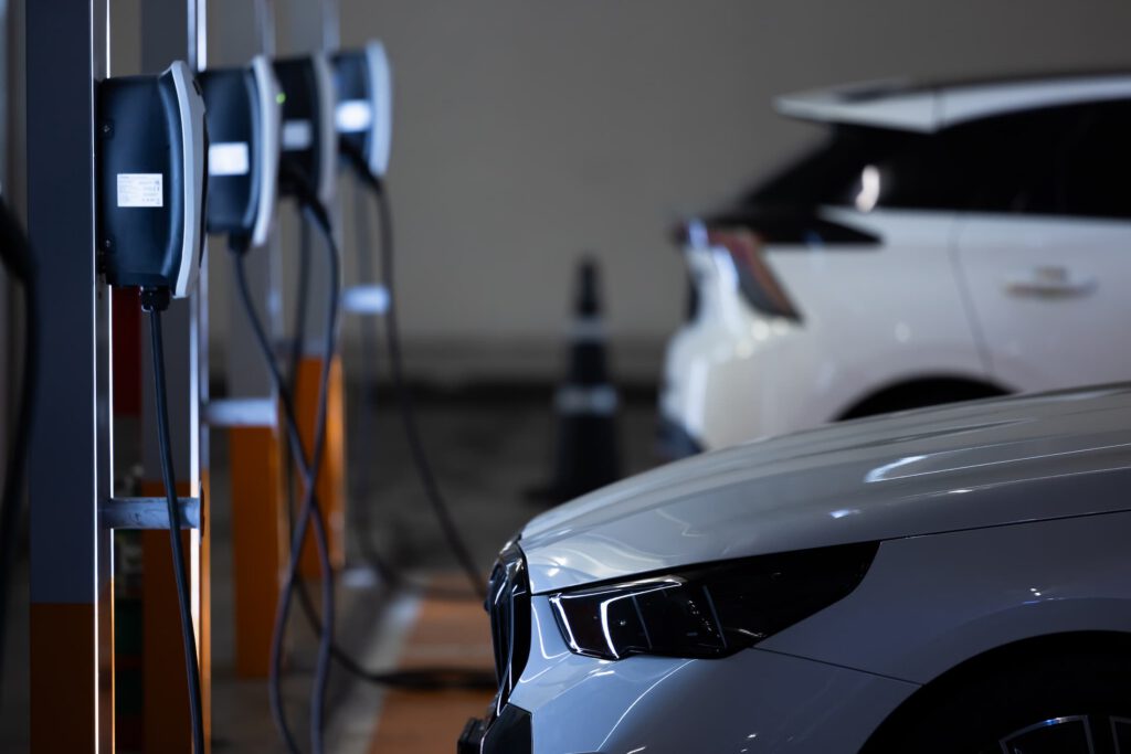 Close-up of two white electric cars parked at charging stations, plugged in, with charging cables visible in an indoor parking area. The focus is on the cars’ front ends and the charging equipment.