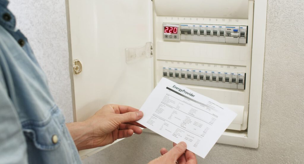 A person holding an energy bill in front of an open electrical panel with switches and a digital display showing "220". The person appears to be checking the bill against the electrical panel.