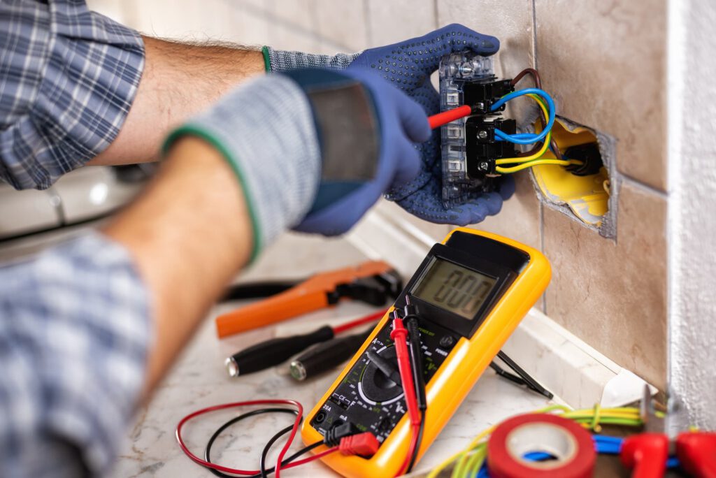 An electrician wearing gloves repairs a wall outlet with colorful wires using tools. A digital multimeter displays zero and various hand tools are visible on a countertop nearby.