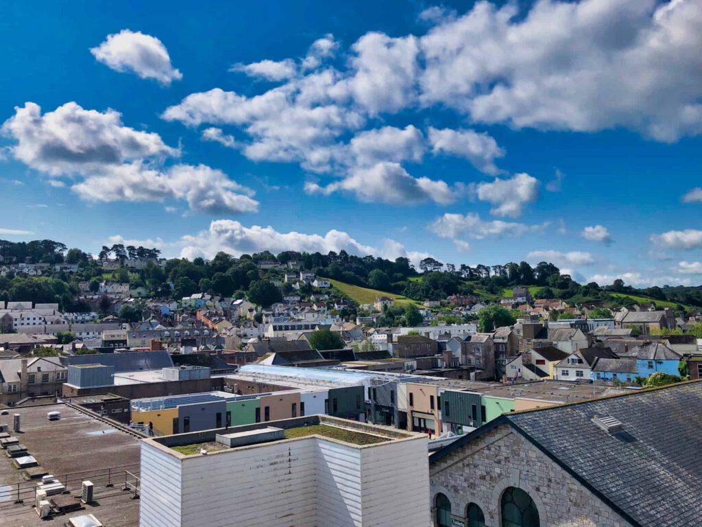 A scenic view of a small town with rows of houses and buildings, surrounded by green hills and trees under a bright blue sky with scattered clouds.