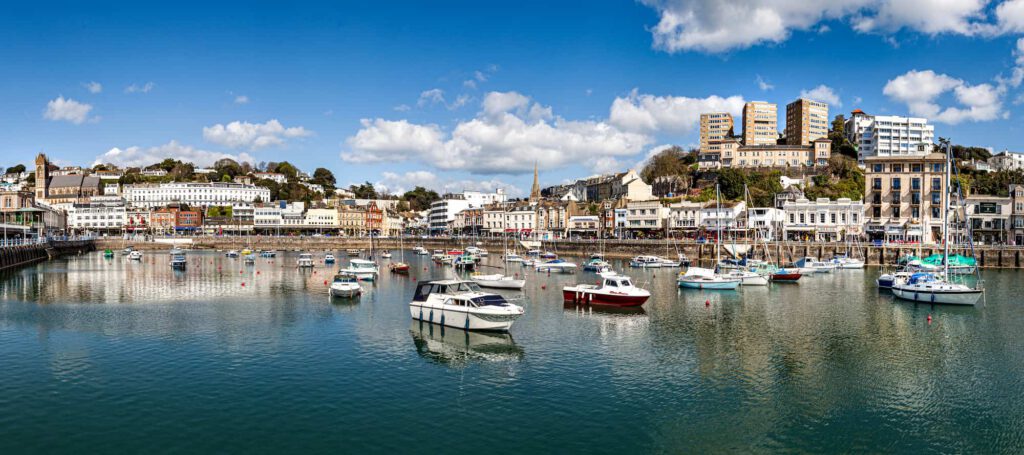 A scenic harbor with various boats floating on calm water, lined by a row of white buildings and taller structures on a hillside under a blue sky with scattered clouds.