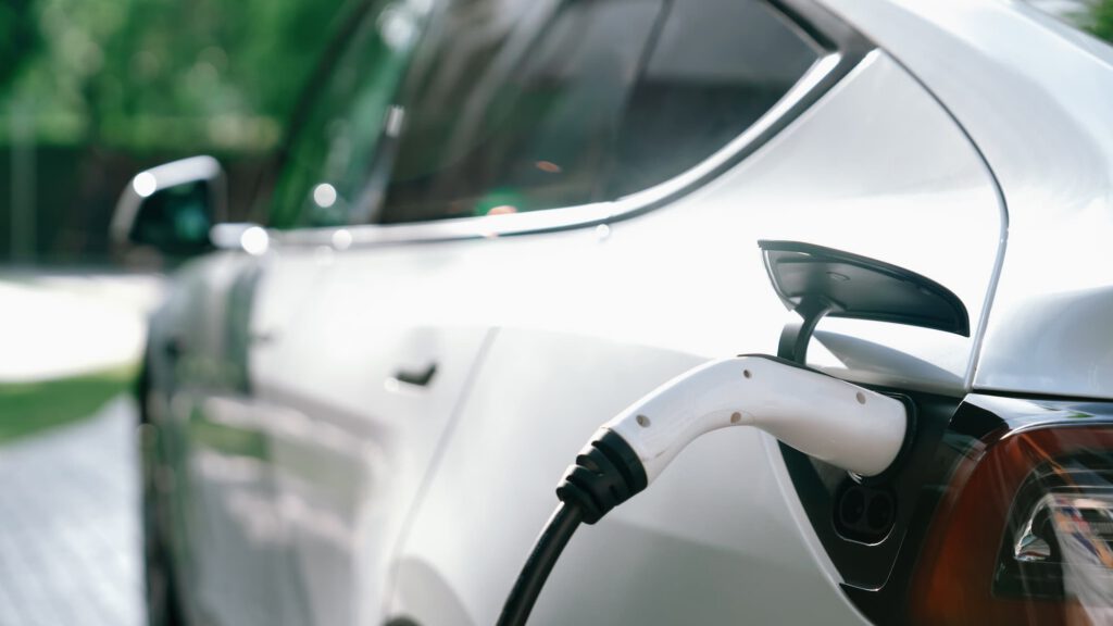 A close-up view of a white electric car being charged, with a charging cable plugged into its port. The background shows greenery and sunlight reflecting off the car's window.